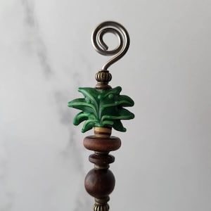 May include: A green glass pineapple charm with brown wooden beads and a silver metal hook. The charm is attached to a metal rod.