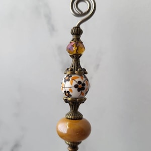 May include: A decorative metal pin with a swirl at the top, a brown and white ceramic bead with a floral pattern, and a brown glass bead.
