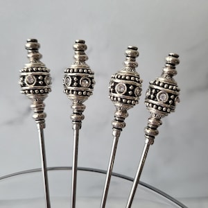 May include: Set of four silver appetizer picks with decorative, beaded tops. Each top features a silver bead with a geometric pattern and clear rhinestones.