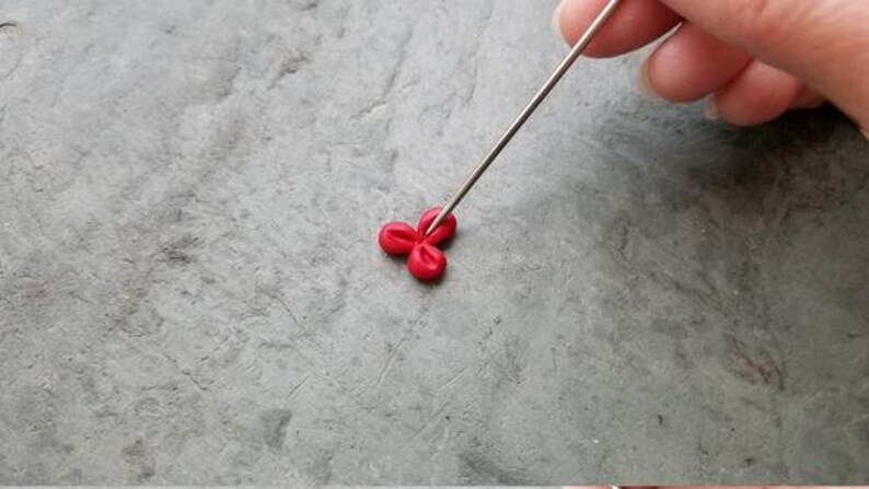 May include: A close-up of a red clay flower being shaped with a metal tool. The flower is made of three petals and is sitting on a gray surface.