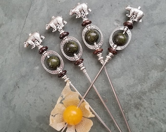 Puede incluir: Cuatro pinchos de plata con cuentas verdes y marrones y remates en forma de oso. Los pinchos están dispuestos en forma de estrella sobre un fondo gris.