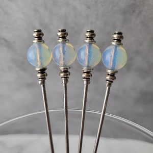 May include: Four silver cocktail sticks with iridescent white opal glass beads on top. The sticks are arranged in a fan shape on a clear glass surface.