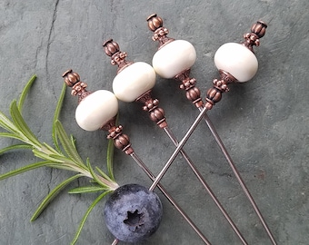 Könnte beinhalten: Vier silberne Cocktailspieße mit weißen Keramik- und kupferfarbenen Aufsätzen. Die Spieße sind auf einer grauen Oberfläche mit einem Rosmarinzweig und einer Blaubeere angeordnet.