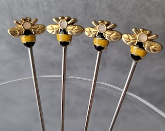 May include: Set of four gold bee cocktail picks with black and yellow stripes and white accents. The picks are made of metal and have a long, thin design.