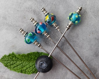 Puede incluir: Cuatro pinchos de cóctel plateados con cuentas de vidrio de colores. Las cuentas son azules, verdes y amarillas con detalles blancos y rojos. Los pinchos están dispuestos en forma de estrella sobre una superficie gris.