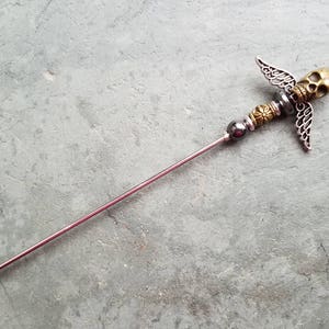 May include: A silver and gold metal hair stick with a skull and wings design. The stick is about 6 inches long.