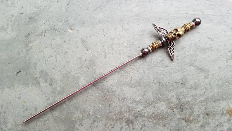 May include: A silver and gold metal hair stick with a skull and wings design. The stick is about 6 inches long.