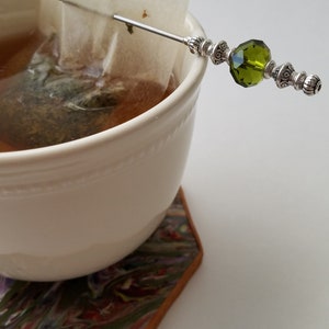 May include: A white ceramic mug with a tea bag and a silver tea stirrer with a green glass bead. The mug is sitting on a colorful coaster.