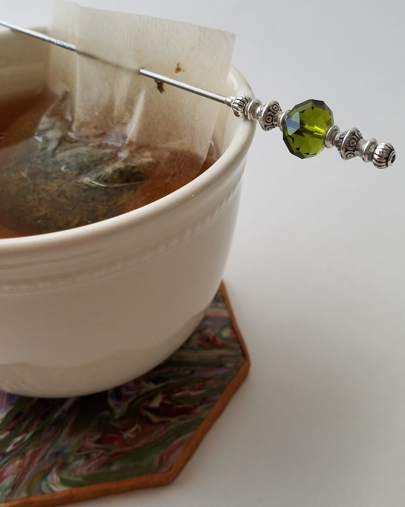 May include: A white ceramic mug with a tea bag and a silver tea stirrer with a green glass bead. The mug is sitting on a colorful coaster.