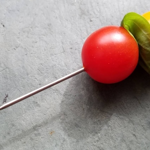May include: A skewer with a red cherry tomato, a green basil leaf, and a yellow cherry tomato.