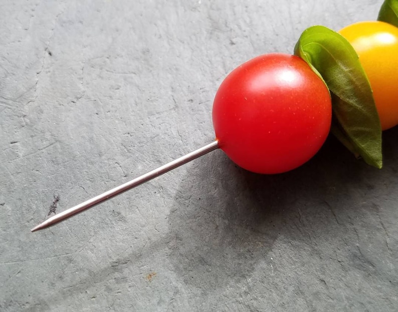 May include: A skewer with a red cherry tomato, a green basil leaf, and a yellow cherry tomato.