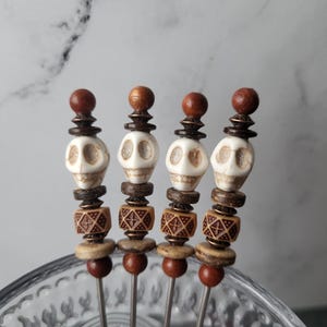 May include: Four metal cocktail picks featuring decorative beads. Each pick is topped with a round brown bead, a white skull-shaped bead, a dark brown spacer, a square brown bead with a geometric pattern, and a light brown disc bead.