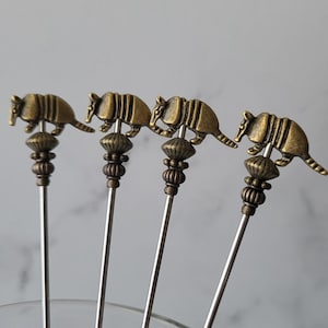 May include: Four gold-toned armadillo shaped cocktail picks with silver metal stems.