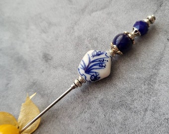 Könnte beinhalten: Eine silberne Nadel mit einer weißen Keramikperle mit blauem Blumenmuster und einer dunkelblauen Glasperle.