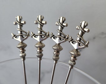 Könnte beinhalten: Vier silberfarbene Cocktailspieße aus Metall, jeder mit einem dekorativen Anker-Aufsatz, der mit einem Seilmuster umwickelt ist. Die Spieße haben einen schlanken, geraden Schaft und eine strukturierte, gestufte Basis unter dem Ankermotiv.