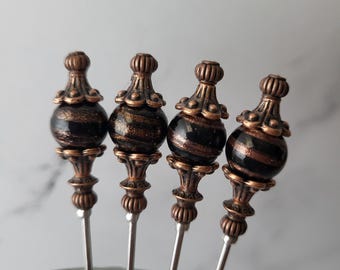 Puede incluir: Juego de cuatro pinchos de cóctel decorativos con cuentas de vidrio rayadas en negro y dorado y tapas de metal de tono cobre.