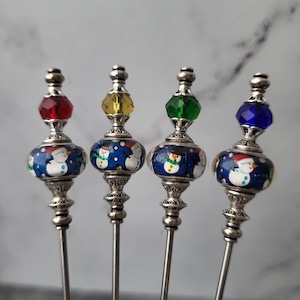 May include: Four decorative cocktail picks with snowman designs. Each pick features a silver stem, a blue and white snowman-decorated bead, and a faceted jewel in red, yellow, green, or blue. The picks are designed for festive occasions.