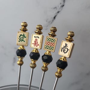May include: Four decorative cocktail picks with gold accents, black faceted beads, and rectangular toppers featuring Chinese characters and designs in red, green, and black. The picks have a silver stem.