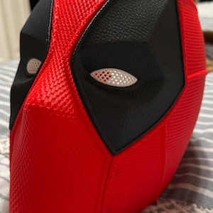 Könnte beinhalten: Eine rot-schwarze Deadpool-Maske. Die Maske hat eine strukturierte rote Oberfläche mit schwarzen Augen- und Gesichtsdetails. Die Augen haben weiße und rote Akzente. Die Maske ist ein Cosplay- oder Kostümartikel.