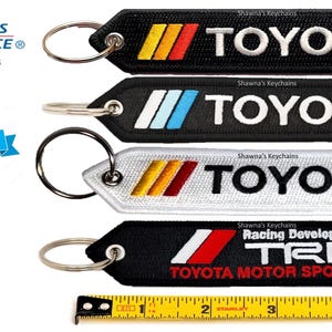 May include: Four black and white keychains with different Toyota logos. The keychains feature a silver metal ring and a black and white fabric tag. The logos include "Toyota", "Toyota Racing Development", and "TRD Toyota Motor Sports".