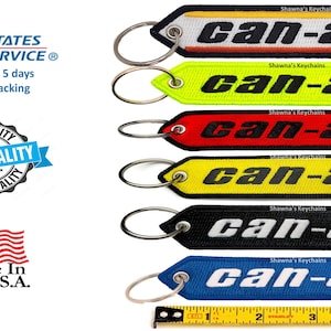 Könnte beinhalten: Sechs verschiedene farbige Schlüsselanhänger mit dem Text "Can-Am" in schwarzer, weißer oder gelber Schrift. Die Schlüsselanhänger sind aus Stoff und haben einen Metallring zum Anbringen von Schlüsseln.