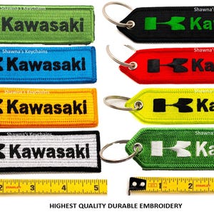 May include: Eight different colored keychains with a Kawasaki logo and a metal ring. The keychains are made of fabric and have a black and white Kawasaki logo. The keychains are all lined up in a row with a ruler underneath.