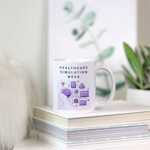 May include: White ceramic mug with the text "HEALTHCARE SIMULATION WEEK" in black. The mug features a purple graphic of a brain, medical symbols, and technology icons. The mug is sitting on a stack of books.