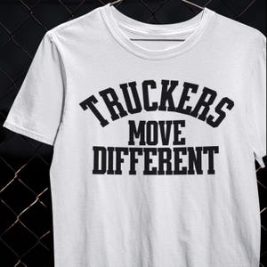 Puede incluir: Camiseta blanca con el texto "TRUCKERS MOVE DIFFERENT" en letras negras y en negrita. La camiseta tiene cuello redondo y mangas cortas. El texto está dispuesto en tres líneas, con "TRUCKERS" arqueado sobre "MOVE DIFFERENT".