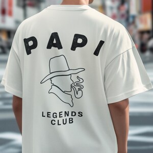 May include: Cream-colored t-shirt with the word "PAPI" in bold black letters across the upper back. Below the text is a black and white graphic of a man in a hat smoking a pipe, with the words "LEGENDS CLUB" underneath.