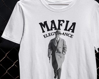 Pode incluir: Camiseta branca com um design gráfico com a palavra "MAFIA" acima de uma imagem de um homem de terno e chapéu, com as palavras "ELEGANCE" e "ANCE" em ambos os lados. A frase "Dressed to kill." é impressa abaixo.