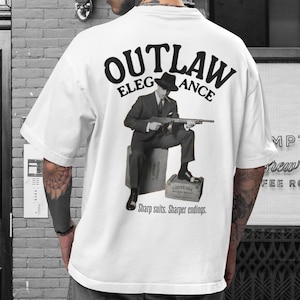 May include: White t-shirt with a graphic on the back featuring a man in a suit holding a gun, with the words "OUTLAW ELEGANCE" above and "Sharp suits. Sharper endings." below. The image is in black and white.