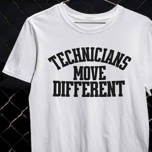 以下が含まれることがあります： 太字の黒いフォントで「TECHNICIANS MOVE DIFFERENT」というテキストがプリントされた白いTシャツ。Tシャツはクルーネックと半袖です。背景は暗く、質感のある表面です。