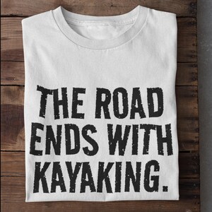 以下が含まれることがあります： 「THE ROAD ENDS WITH KAYAKING.」というテキストが太字の黒い文字で書かれた白いTシャツ。Tシャツは折りたたまれ、木製の表面に置かれています。Tシャツはクルーネックです。