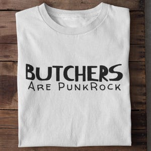 May include: White t-shirt with the black text "BUTCHERS ARE PUNK ROCK" on a wooden surface. The shirt is folded, showing the crew neck and the bold, handwritten-style lettering. A casual, statement tee.