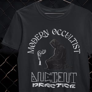 May include: Black t-shirt with a graphic featuring a hooded figure and the words "MODERN OCCULTIST" and "ANCIENT PRACTICE" in white. The design includes a stylized flame and a seated figure.