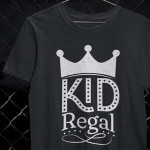 Könnte beinhalten: Schwarzes T-Shirt mit einem weißen Kronen-Design und den Worten "KID Regal". Der Text ist in einer fetten, stilisierten Schriftart mit gepunkteten Akzenten gehalten. Das Shirt besteht aus einem weichen Material.