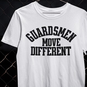 以下が含まれることがあります： 太字の黒いフォントで「GUARDSMEN MOVE DIFFERENT」というテキストがプリントされた白いTシャツ。Tシャツはクルーネックと半袖です。テキストは3行に配置され、「GUARDSMEN」は上部にアーチ状になっています。