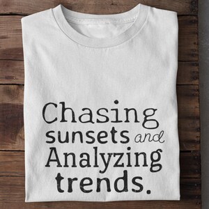 Puede incluir: Camiseta blanca con el texto "Chasing sunsets and Analyzing trends." La camiseta está hecha de un material suave y está doblada sobre una superficie de madera. El texto está en negrita negra.