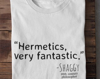 May include: White t-shirt with the quote "Hermetics, very fantastic." attributed to Shaggy, a 20th-century philosopher. The text is in black, and the shirt is folded on a wooden surface.