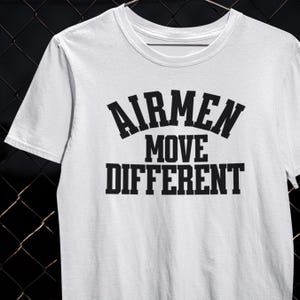以下が含まれることがあります： 白のクルーネックTシャツ。前面に黒の太字で「AIRMEN MOVE DIFFERENT」の文字がプリントされています。柔らかい素材でできており、半袖です。デザインはシャツの中央に配置されています。