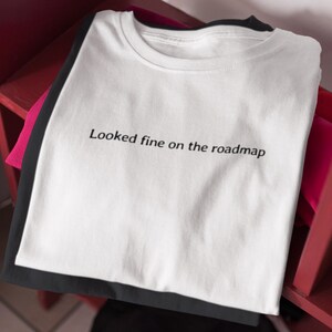 May include: A stack of folded t-shirts in white, black, and pink. The white t-shirt on top has the phrase "Looked fine on the roadmap" printed in black, centered on the front. The shirts are stacked on a red surface.