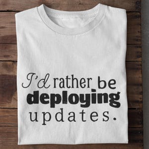 May include: White t-shirt with the text "I'd rather be deploying updates." The text is in a mix of cursive and bold fonts. The shirt is folded and placed on a wooden surface.