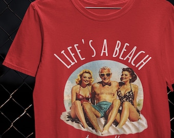 May include: Red t-shirt featuring the phrase "LIFE'S A BEACH" above a retro illustration of a man and two women on a beach. The words "stay salty" are printed below the illustration.