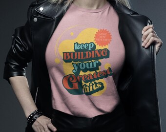 May include: A pink t-shirt with the text "Keep Building Your Greatest Hits" in a retro font. The shirt has a yellow and orange design. The person is wearing a black leather jacket and a black bracelet.