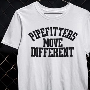 Può includere: T-shirt bianca con la scritta nera "PIPEFITTERS MOVE DIFFERENT". Il testo è in grassetto e disposto su tre righe. La maglietta ha un girocollo e maniche corte, adatta per un look casual.
