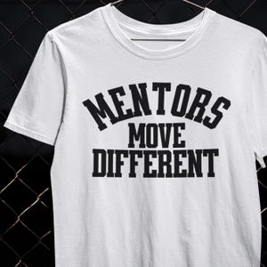 Puede incluir: Camiseta blanca con el texto "MENTORS MOVE DIFFERENT" en letras negras y en negrita. La camiseta tiene cuello redondo y mangas cortas. El diseño está centrado en la parte delantera.