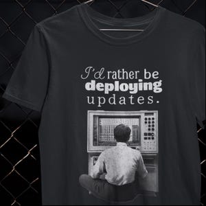 Può includere: T-shirt nero con la scritta "I'd rather be deploying updates." Sotto il testo c'è una grafica di un computer vintage, che mostra una persona seduta davanti a un computer. La maglietta è girocollo.