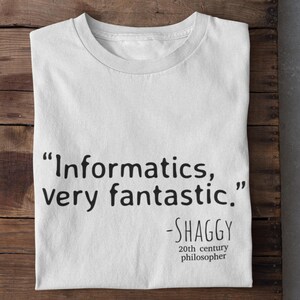 以下が含まれることがあります： 「Informatics, very fantastic.」という引用句が黒いテキストで書かれた白いTシャツ。20世紀の哲学者であるシャギーによるもの。木製の表面に折りたたまれています。