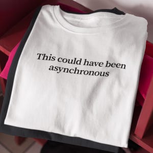 May include: White t-shirt with the text "This could have been asynchronous" in black. The shirt is folded and stacked with other shirts, with a black shirt visible underneath. The shirts are on a red shelf.