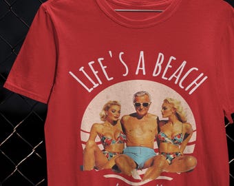 May include: Red t-shirt featuring the phrase "LIFE'S A BEACH" above a retro graphic of three people on a beach. The graphic shows a man in blue swim shorts and two women in floral bikinis. The words "stay salty" are below the graphic.